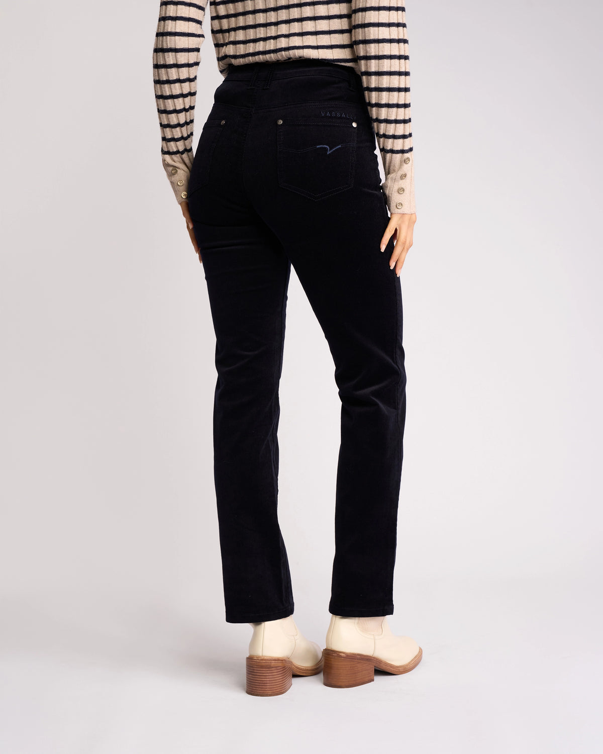 A person wearing Vassalli's Arch Cord Pant in Midnight stands facing away, paired with a long-sleeve striped shirt and light heeled boots, showcasing the chic winter look against a plain background.