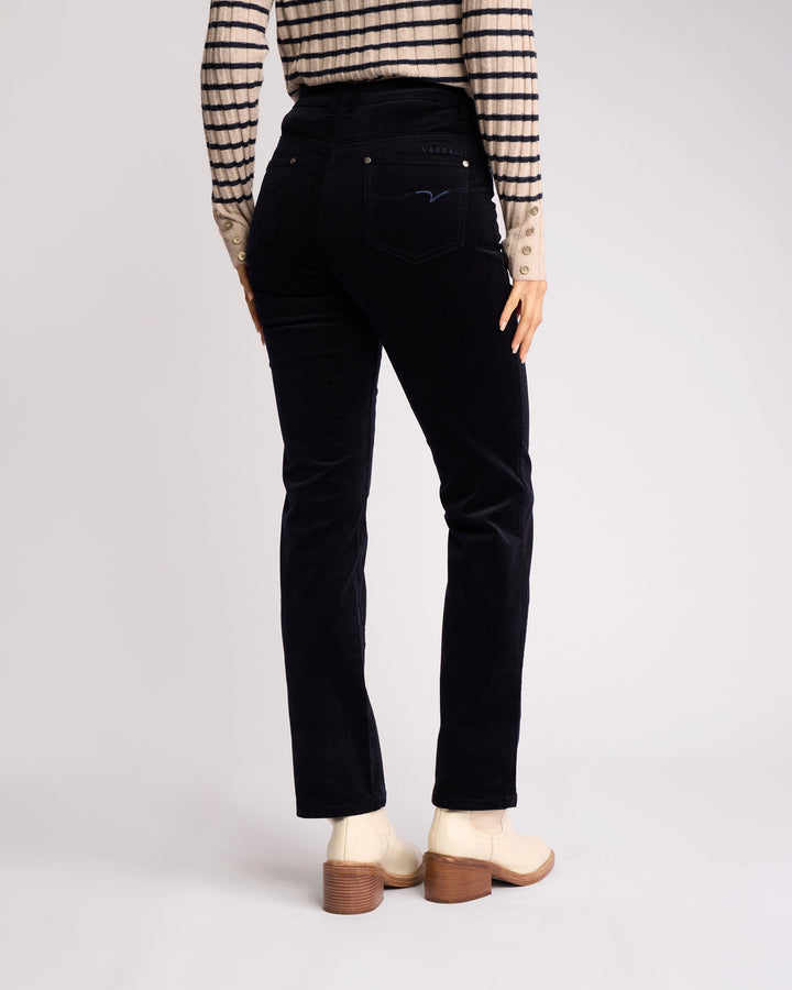 A person wearing Vassalli's Arch Cord Pant in Midnight stands facing away, paired with a long-sleeve striped shirt and light heeled boots, showcasing the chic winter look against a plain background.