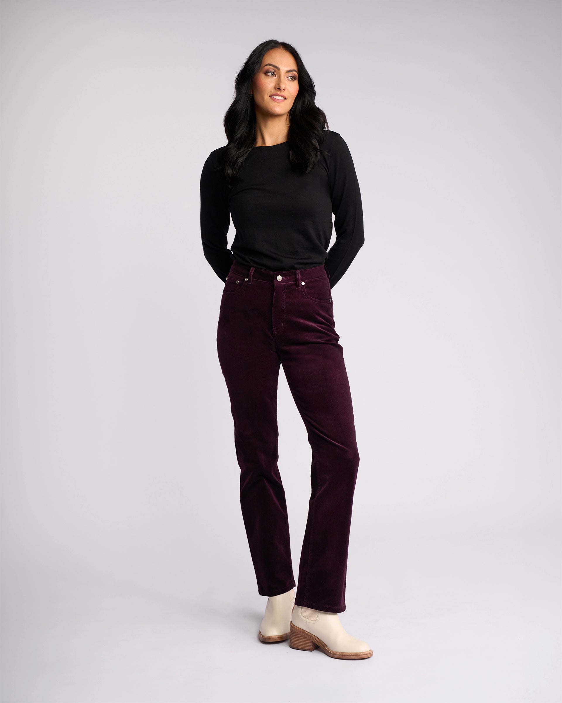 A woman with long dark hair stands against a light gray background, wearing a black long-sleeve top, Vassalli Arch Cord Pant in Mulberry, and light beige ankle boots—an elegant winter wardrobe choice. She looks slightly to the side and smiles softly.