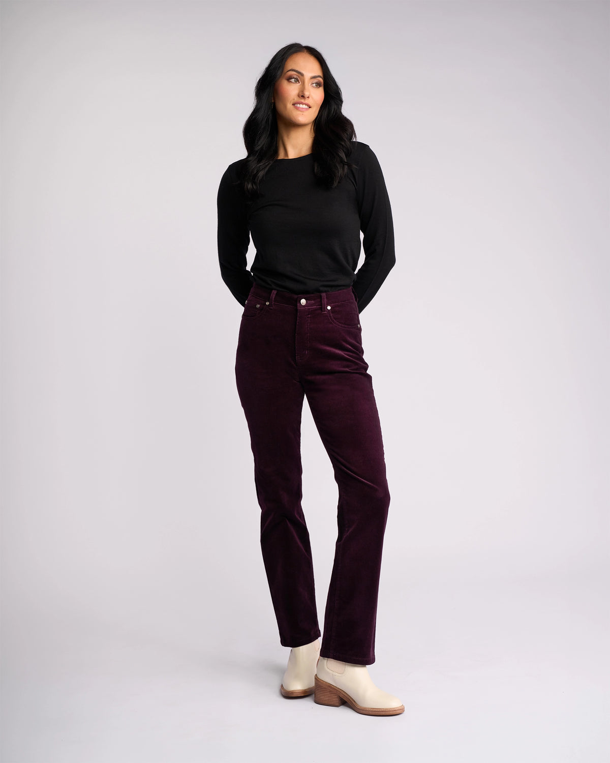 A woman with long dark hair stands against a light gray background, wearing a black long-sleeve top, Vassalli Arch Cord Pant in Mulberry, and light beige ankle boots—an elegant winter wardrobe choice. She looks slightly to the side and smiles softly.