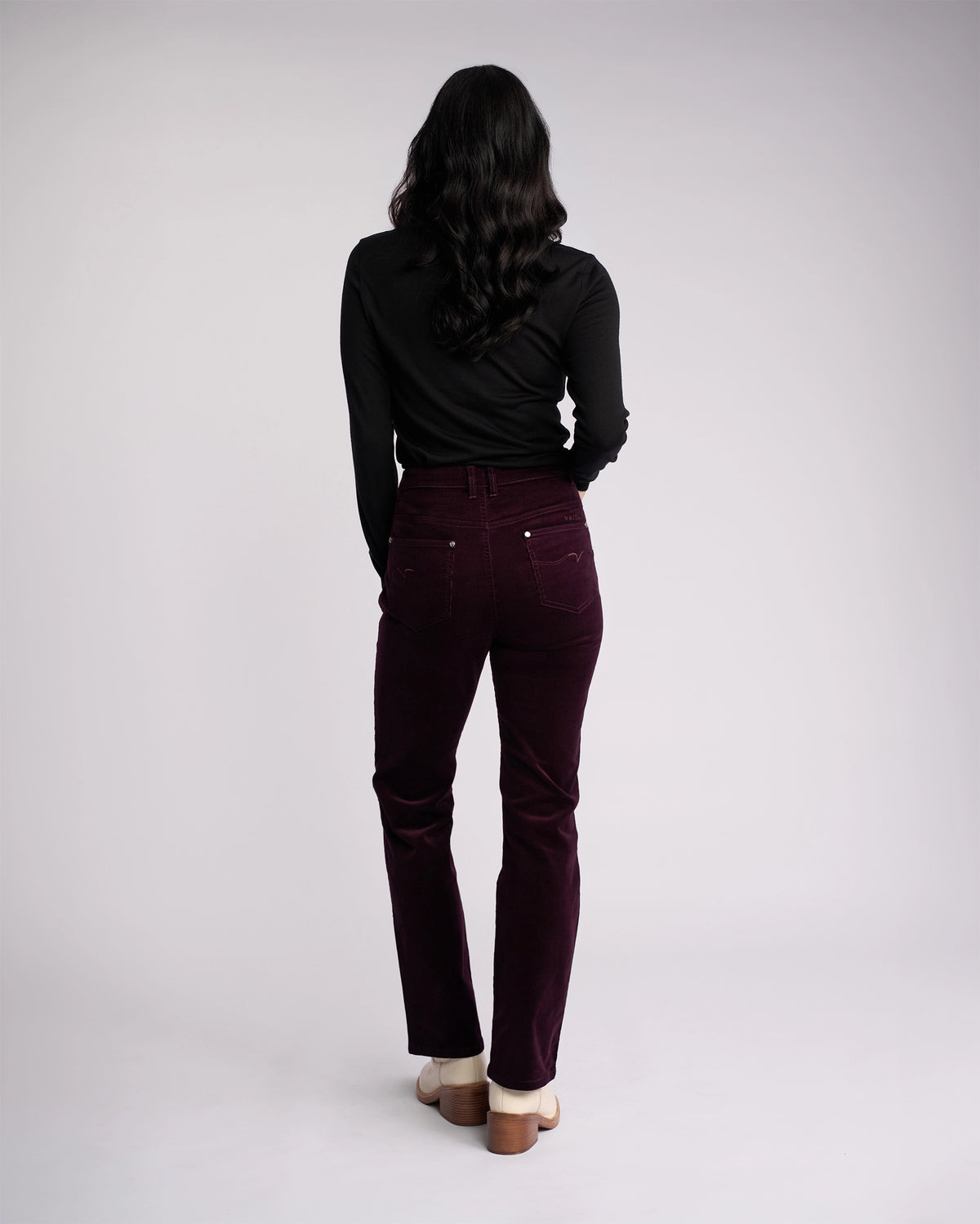 A person with long dark hair, in a black long-sleeve top, Vassalli Arch Cord Pant - Mulberry, and cream heeled shoes stands facing away from the camera against a light background—perfect for a winter wardrobe.