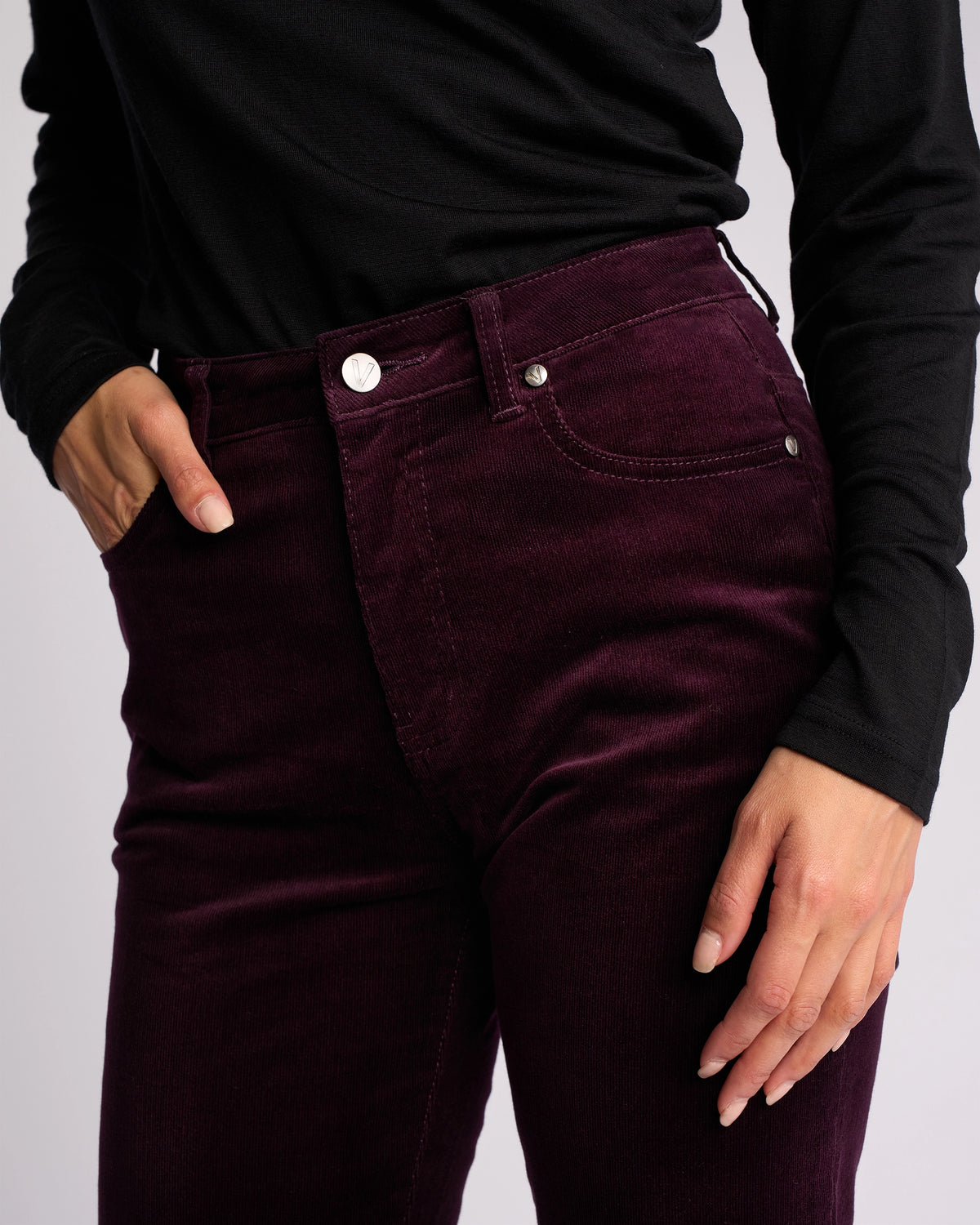 A person wears a black long-sleeve top and high-waisted Vassalli Arch Cord Pant in Mulberry, pinwale cord, with one hand in the pocket—an ideal winter look shown from mid-torso to thighs.