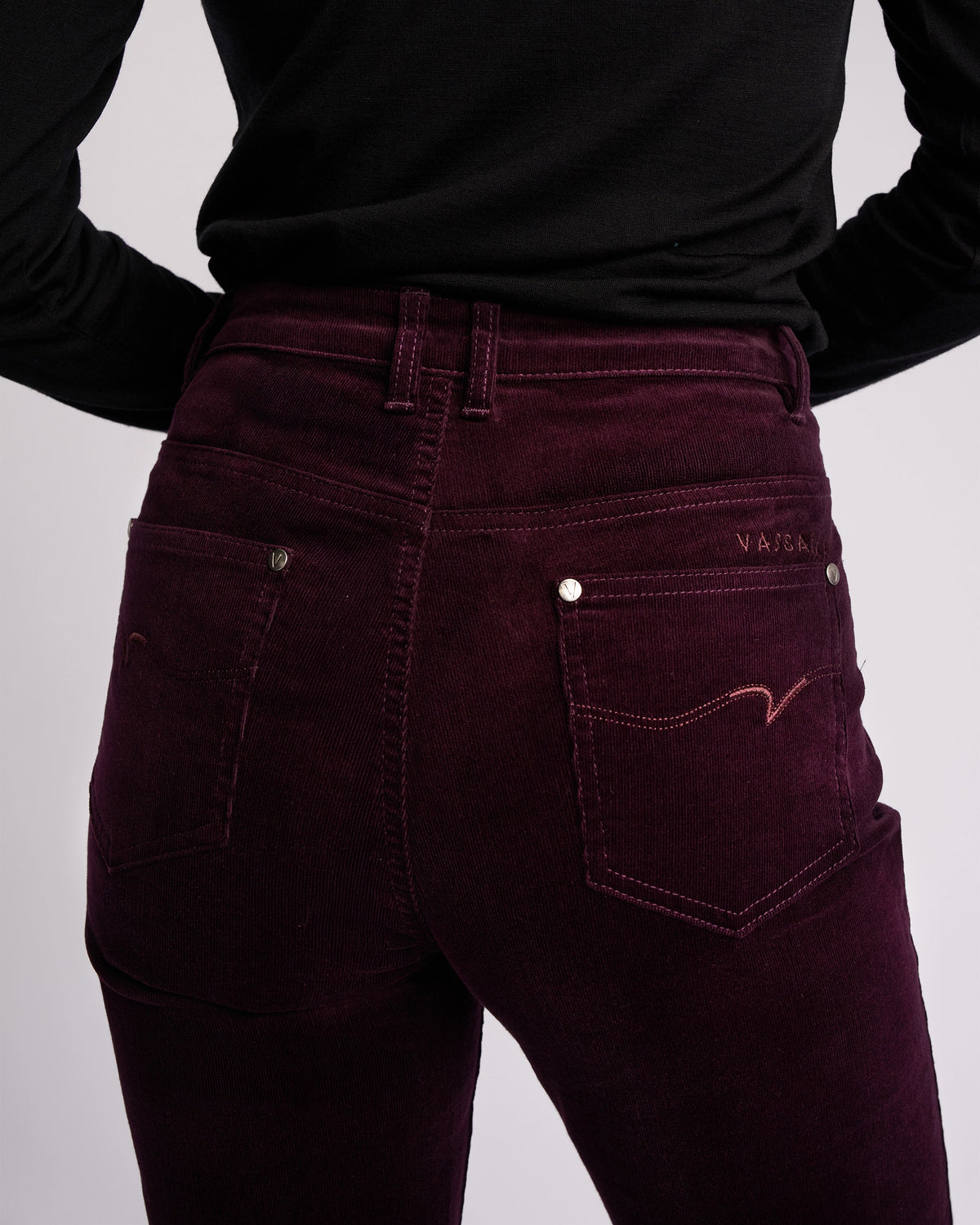 A person wearing a black long-sleeve top and Vassalli Arch Cord Pant in Mulberry is shown from the back against a plain light background—perfect for elevating your winter wardrobe.