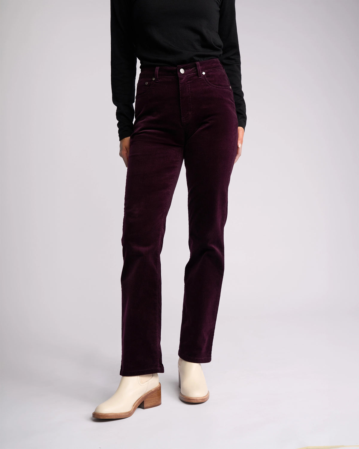 A person models the Vassalli Arch Cord Pant in Mulberry with a long-sleeve black top and off-white heeled ankle boots, standing against a plain light background—a chic winter look with their head out of frame.