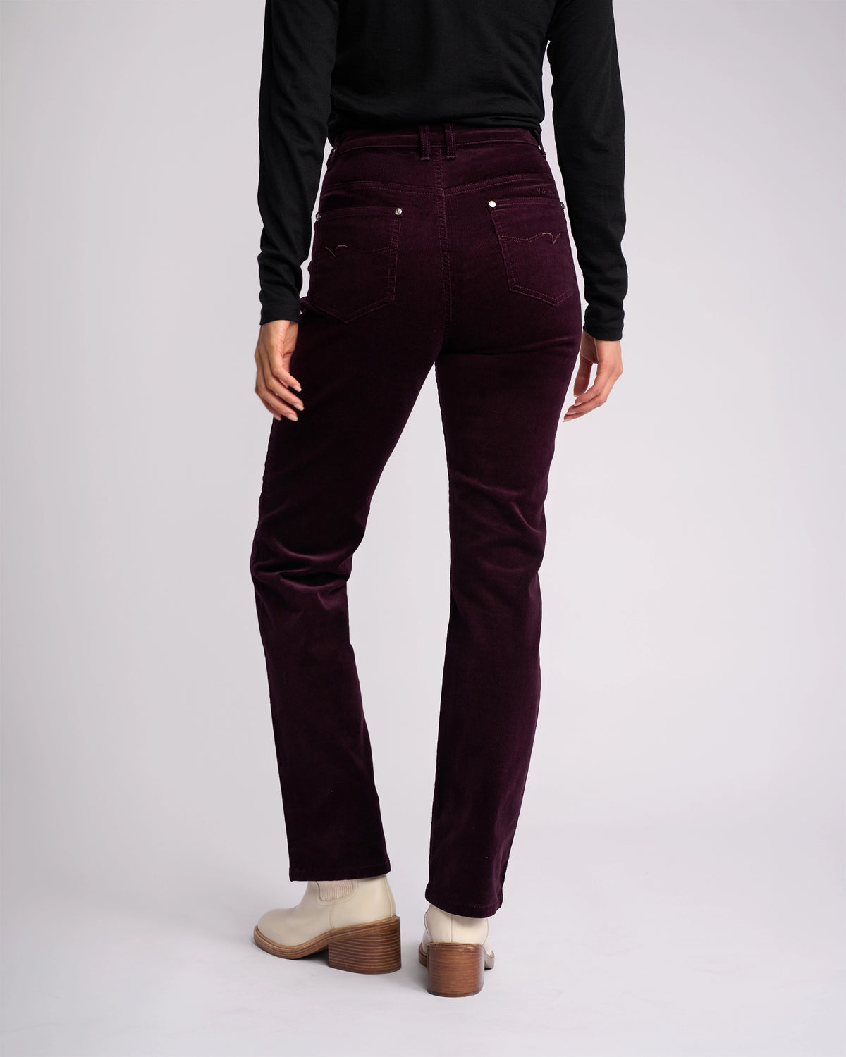 A person faces away from the camera against a plain light background, wearing a black long-sleeve top, cream ankle boots, and Vassalli’s Arch Cord Pant in Mulberry—a stylish winter wardrobe look.