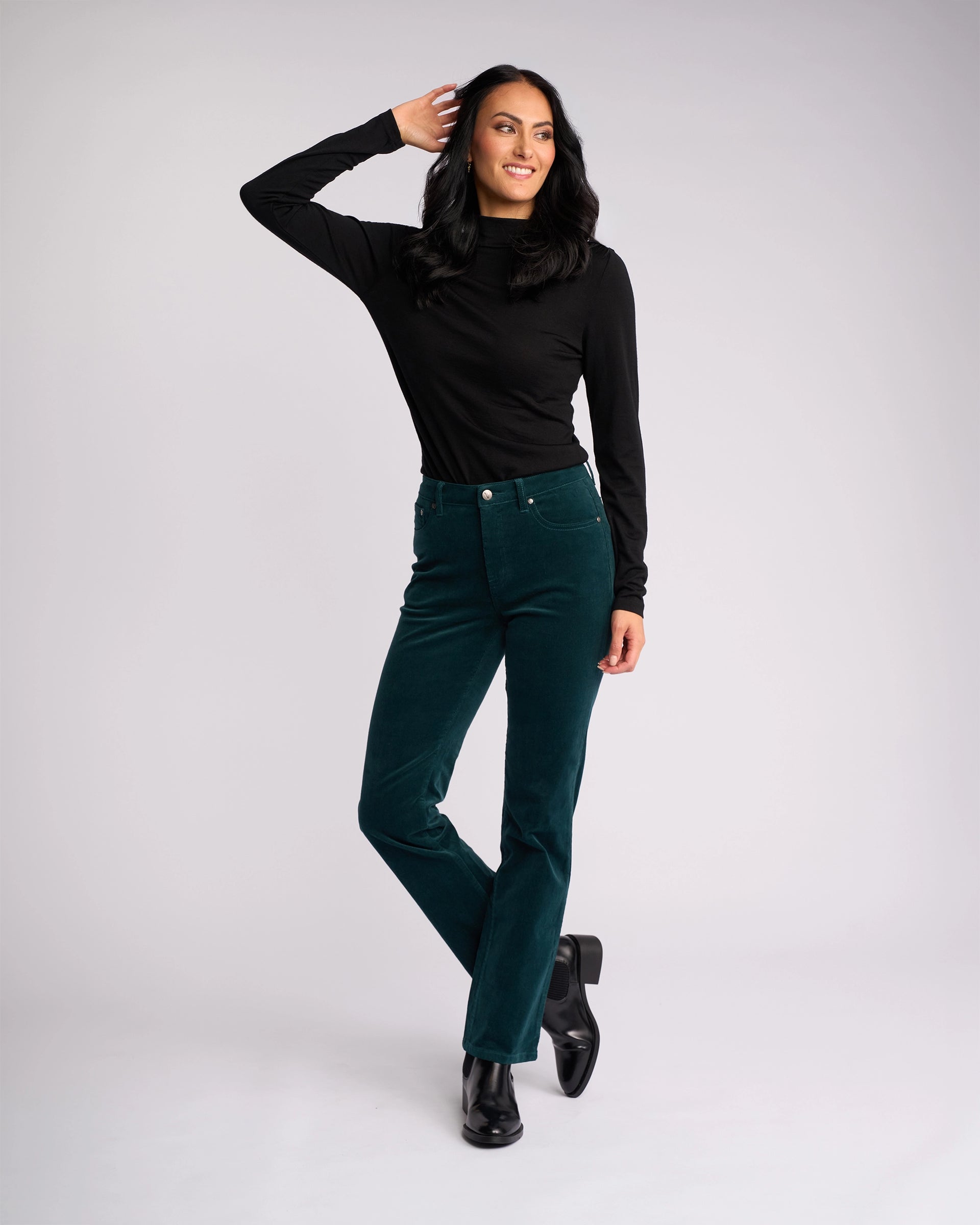 A woman with long dark hair smiles in a sleek winter look—she wears a black turtleneck, Vassalli's Arch Cord Pant in Peacock, and black ankle boots, one hand touching her head and one leg crossed before a light background.