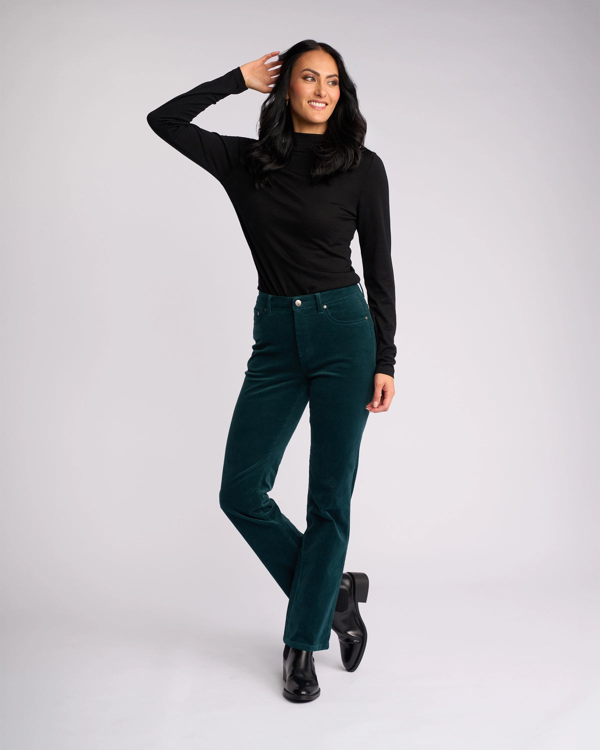 A woman with long dark hair smiles in a sleek winter look—she wears a black turtleneck, Vassalli's Arch Cord Pant in Peacock, and black ankle boots, one hand touching her head and one leg crossed before a light background.