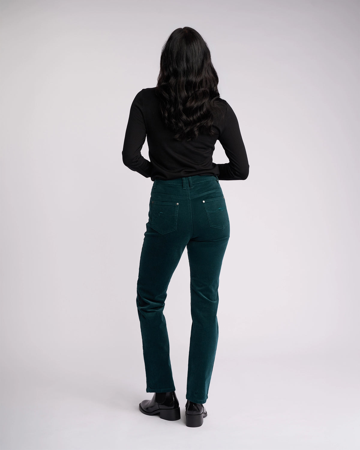 A person with long, wavy dark hair faces away, wearing a black long-sleeve top, Vassalli's Arch Cord Pant in Peacock, and black shoes—an ideal winter look—set against a plain light gray background.