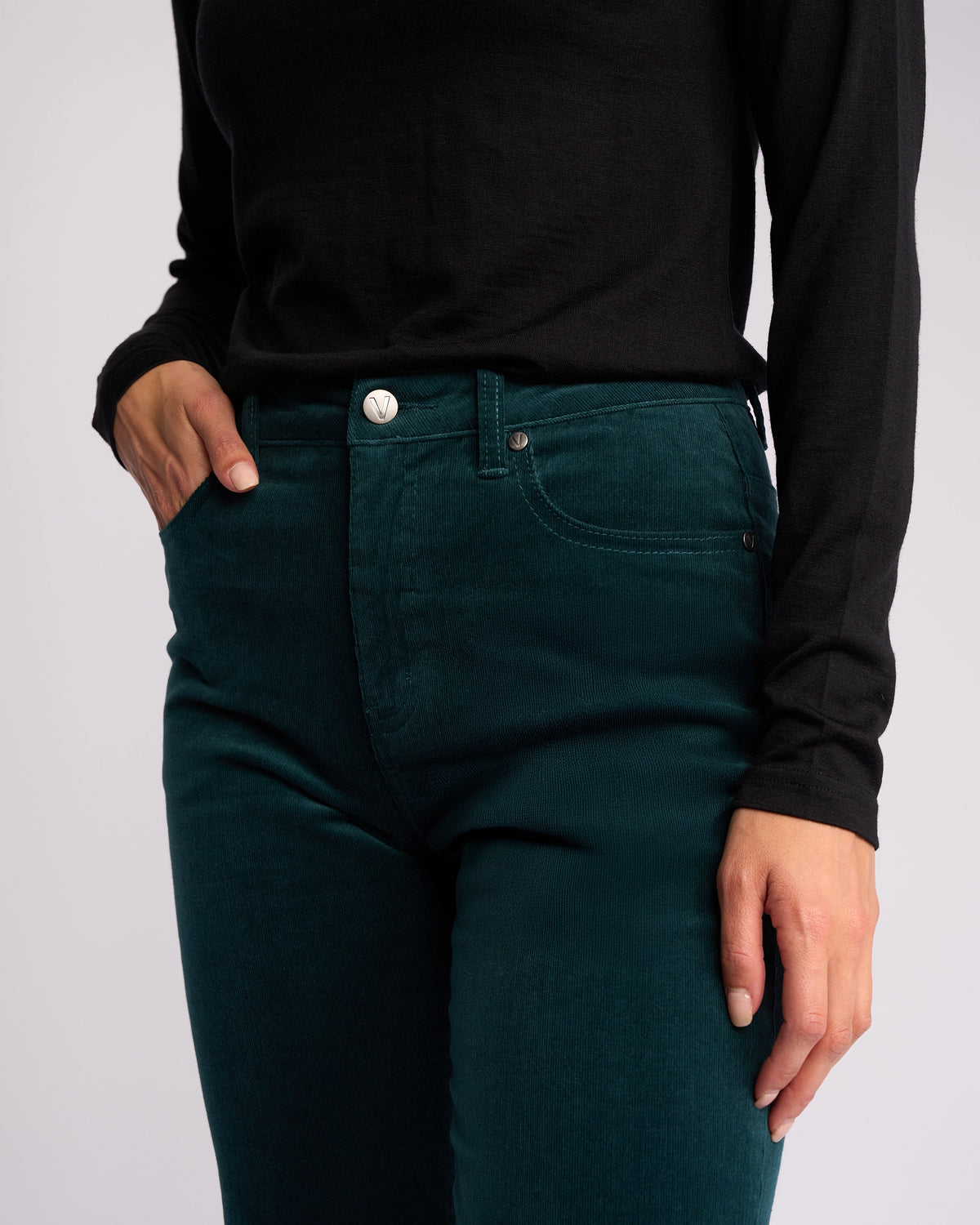 A person wearing a black long-sleeve shirt and Vassalli’s Arch Cord Pant in Peacock stands with one hand in their pocket against a plain background, showcasing this versatile winter wardrobe staple.