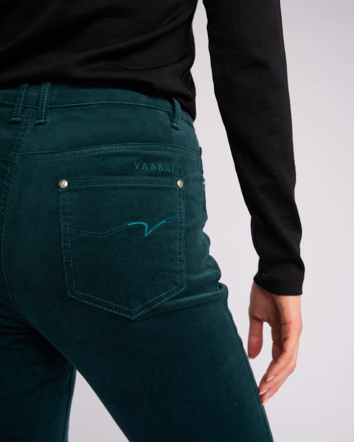 Person wearing Vassalli Arch Cord Pant in Peacock, with straight legs and a back pocket featuring decorative stitching and the Vassalli logo. Styled with a black long-sleeve shirt—ideal for winter wardrobes. Shown from the back, waist down.