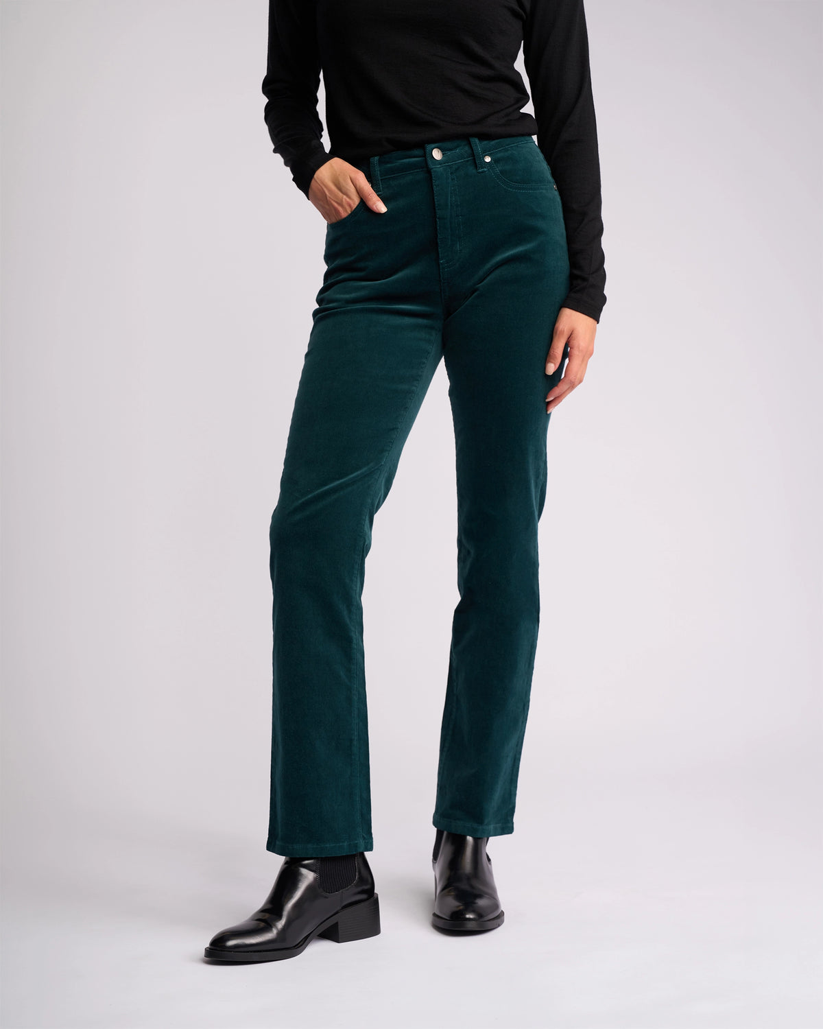 A person in a black long-sleeve top, Vassalli’s Arch Cord Pant - Peacock, and black ankle boots stands against a plain light background with their left hand in their pocket, highlighting a chic winter look.