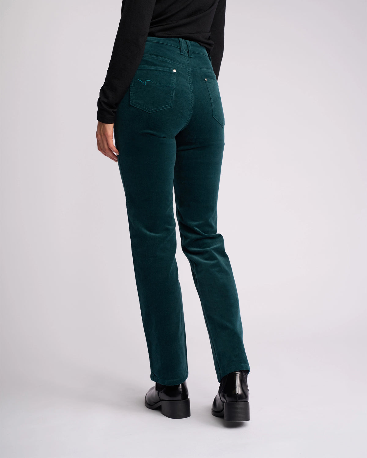 Wearing Vassalli's Arch Cord Pant in Peacock, a person pairs it with a black long-sleeve top and black shoes, standing with their back to the camera against a light background—ideal for refreshing your winter wardrobe.