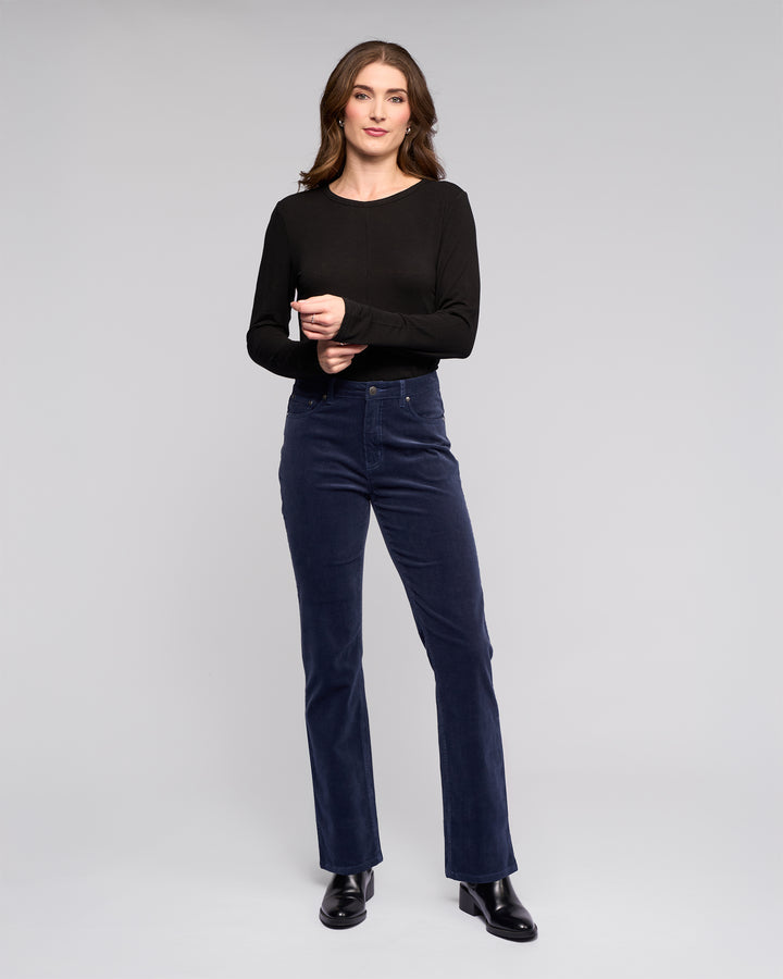A woman stands against a plain light gray background, wearing Vassalli’s Arch Cord Pant in Slate, a long-sleeved black top, and black ankle boots. She has long brown hair and smiles gently with her hands clasped.