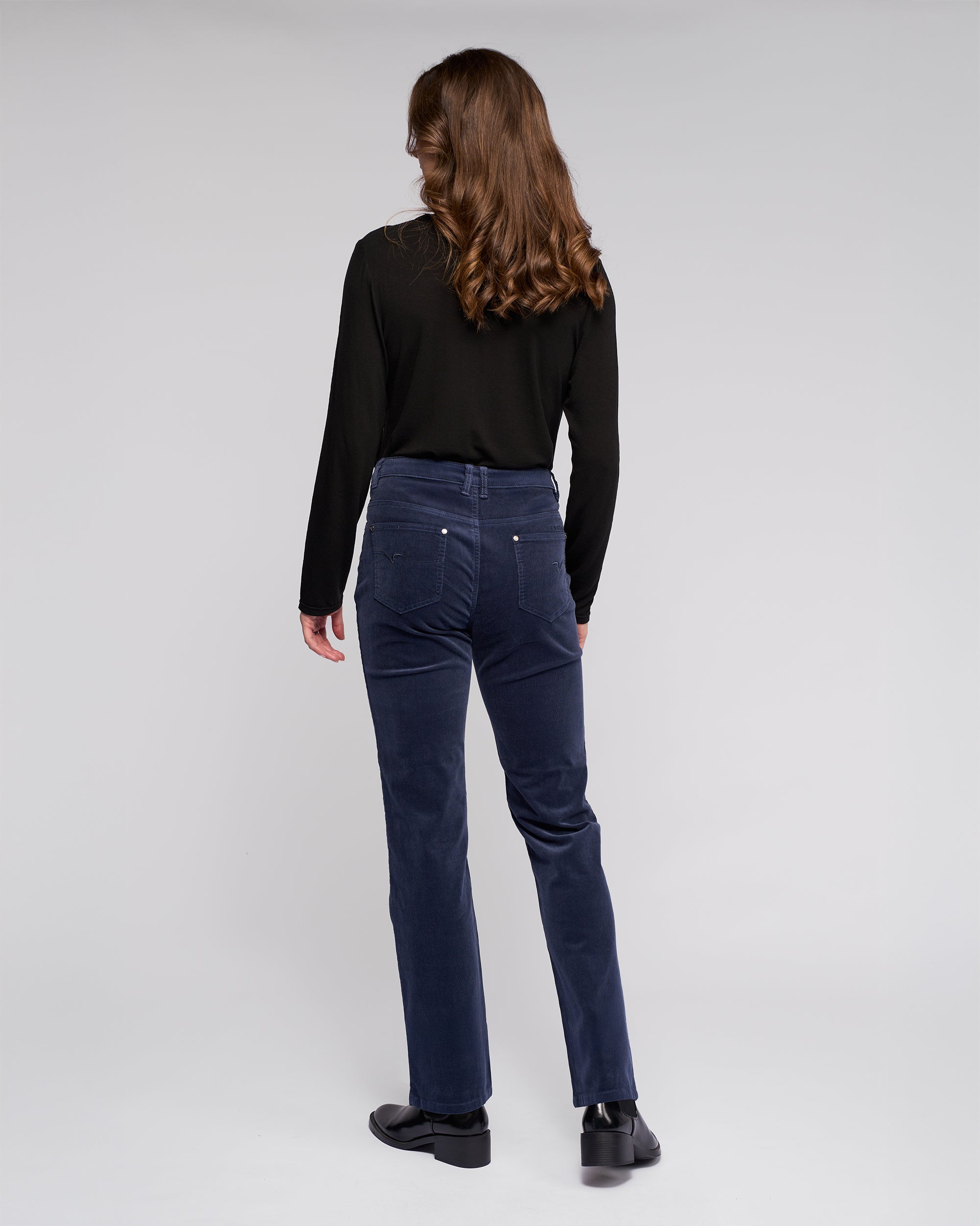 A person with long brown hair, wearing a black long-sleeve top, Vassalli Arch Cord Pant in Slate, and black shoes stands facing away against a plain light gray background.