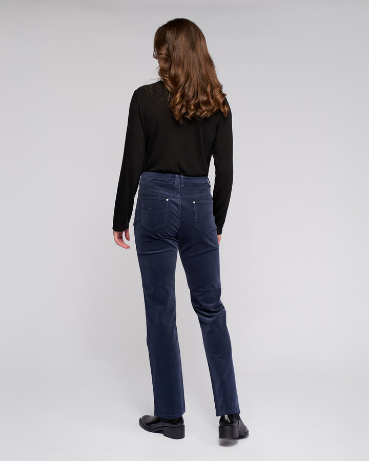 A person with long brown hair, wearing a black long-sleeve top, Vassalli Arch Cord Pant in Slate, and black shoes stands facing away against a plain light gray background.