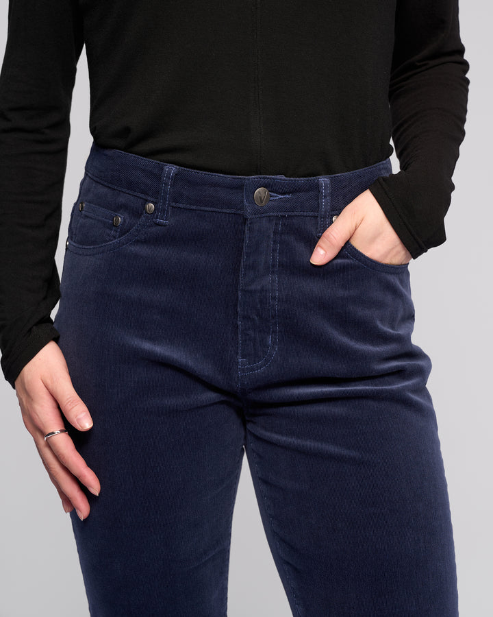 A person wears a black long-sleeve top and Vassalli Arch Cord Pant in Slate, standing with one hand in their pocket. The image is cropped to show only the torso and upper legs.