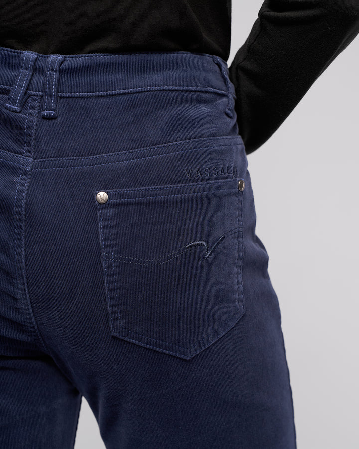 A person is shown from the back wearing Vassalli's Arch Cord Pant in Slate, paired with a black top. The focus is on the waistband and back pocket, which features embroidered details and the Vassalli brand name above the pocket.