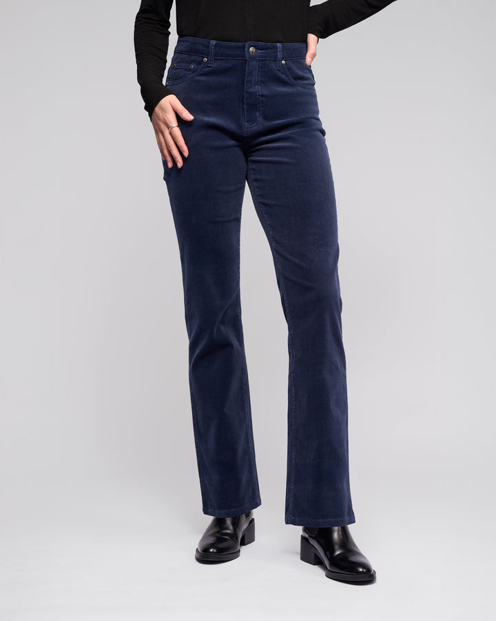 A person models Vassalli's Arch Cord Pant in Slate, paired with a black long-sleeve top and black heeled boots. The photo, cropped from the shoulders down, shows them standing against a plain light background.