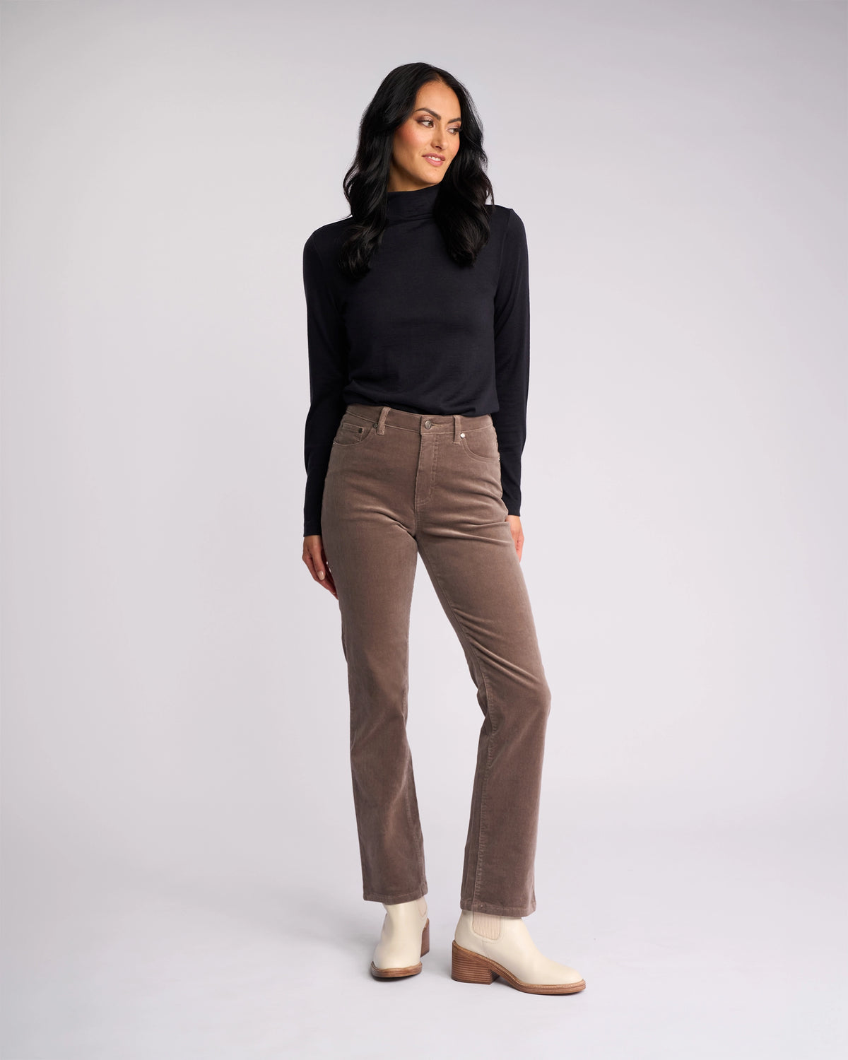 Woman with long dark hair stands against a plain background, wearing a black long-sleeve top, white ankle boots, and Vassalli's Arch Cord Pant in Taupe—an essential for any winter wardrobe. She looks slightly to the side with a relaxed expression.