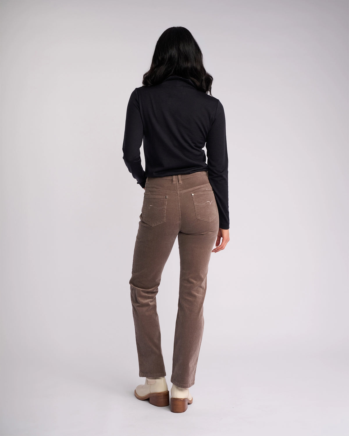 A person with long dark hair, in a black long-sleeve top, Vassalli Arch Cord Pant - Taupe, and light shoes stands facing away from the camera against a plain light background—a timeless winter staple.