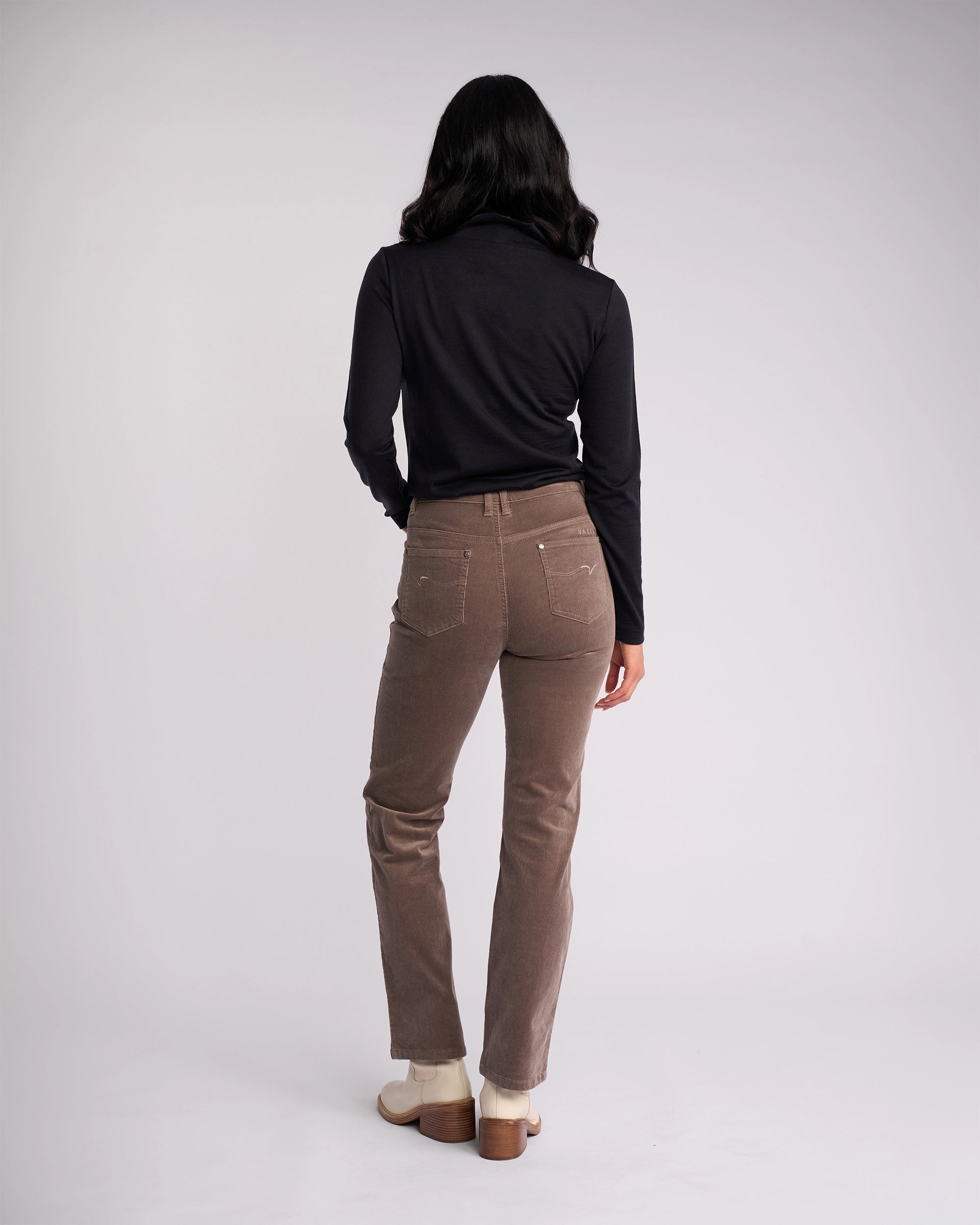 A person with long dark hair, in a black long-sleeve top, Vassalli Arch Cord Pant - Taupe, and light shoes stands facing away from the camera against a plain light background—a timeless winter staple.