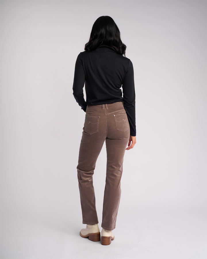 A person with long dark hair, in a black long-sleeve top, Vassalli Arch Cord Pant - Taupe, and light shoes stands facing away from the camera against a plain light background—a timeless winter staple.