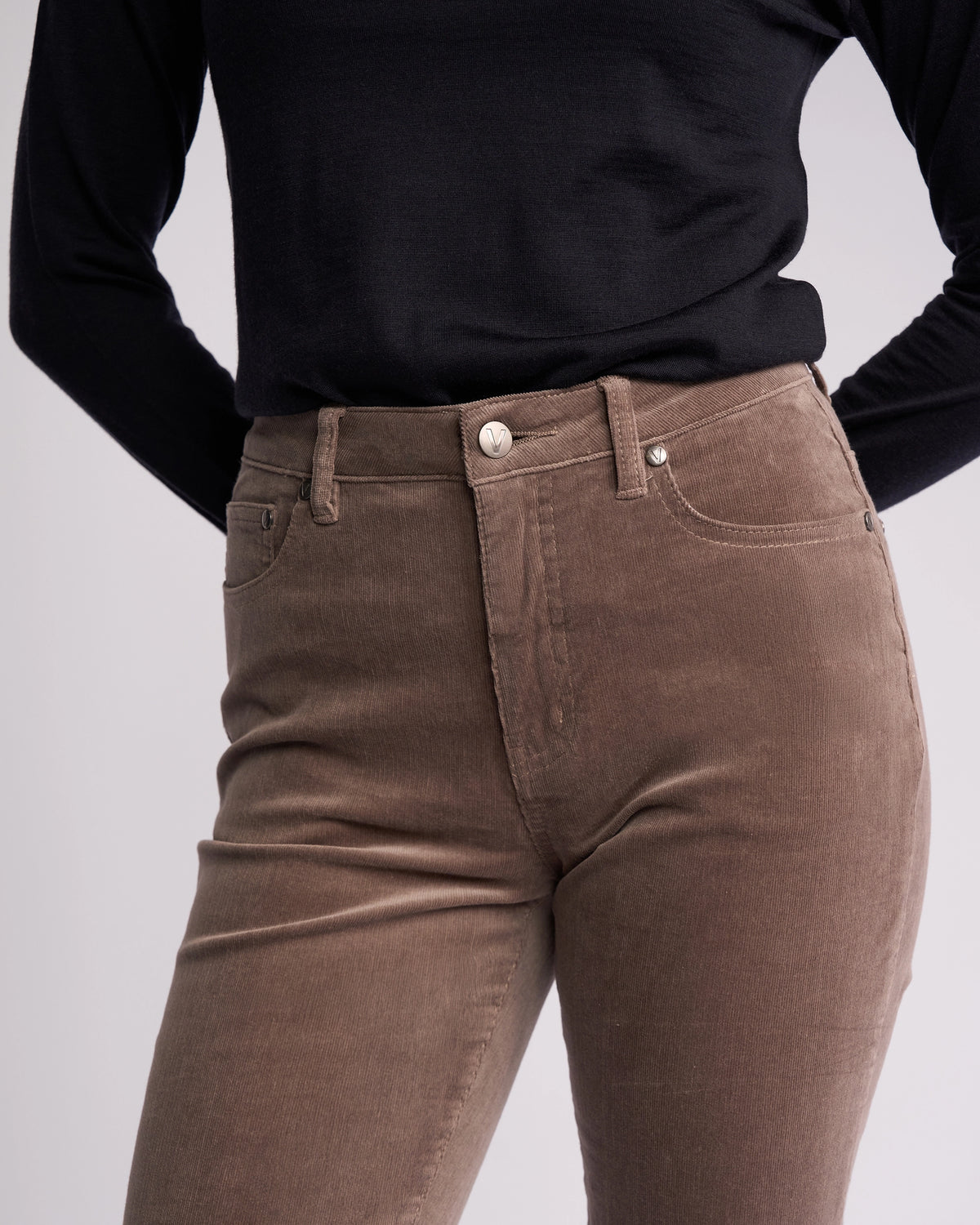 A person wears a black long-sleeve top and Vassalli’s Arch Cord Pant in Taupe, shown from shoulders to mid-thigh with hands behind their back against a plain background—perfect for your winter wardrobe.