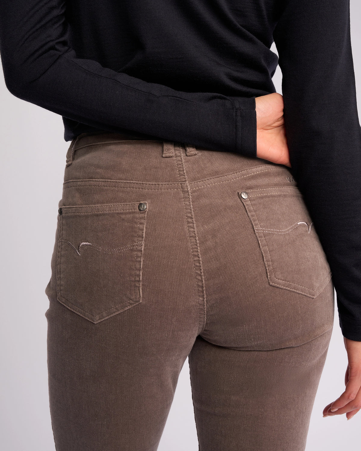 Wearing Vassalli's Arch Cord Pant - Taupe—straight-leg corduroy ideal for winter—and a black long-sleeve shirt, a person stands with one hand behind their back, highlighting the pants' back pockets.