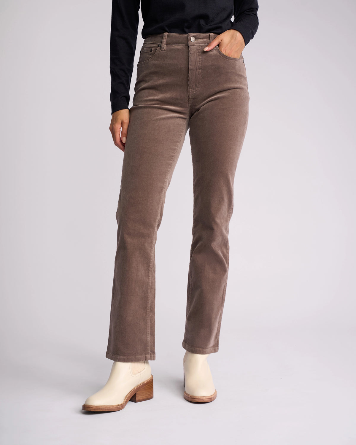 A person wearing the Vassalli Arch Cord Pant in taupe, paired with a black long-sleeve top and beige ankle boots, stands with one hand in their pocket—an effortlessly cool look for any winter wardrobe.