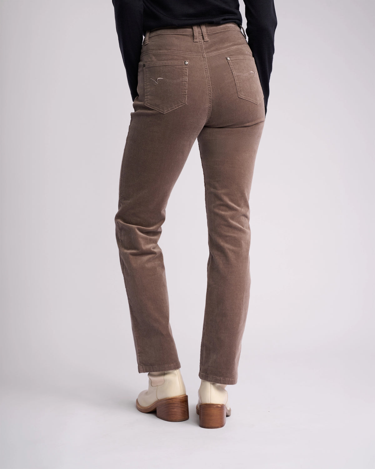 A person stands facing away, wearing Vassalli Arch Cord Pant in taupe with a black top and cream heeled shoes against a light background—an effortless addition to your winter wardrobe.