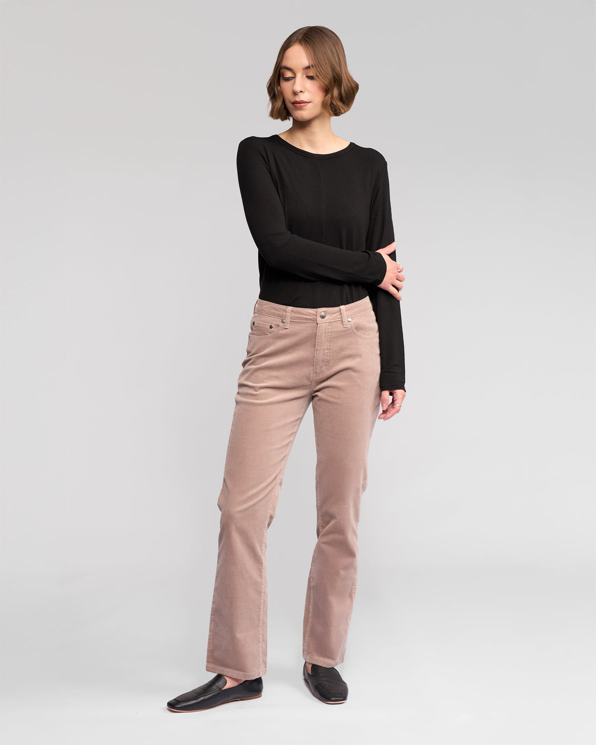 A woman with short brown hair stands against a plain background, wearing a black long-sleeve top, Vassalli Arch Cord Pant in Twig, and black shoes. She crosses one arm over her body and looks slightly to the side.