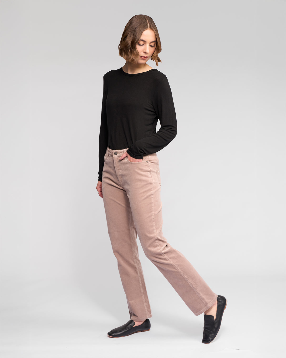 A person with chin-length brown hair wears a long-sleeve black top, Vassalli Arch Cord Pant - Twig in light pink, and black loafers, standing against a plain light background and looking down with one leg slightly bent.