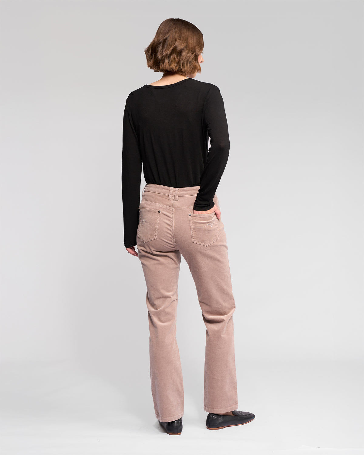 A person with short brown hair, wearing a black long-sleeve top, Vassalli Arch Cord Pant - Twig, and black shoes stands facing away from the camera against a plain light background.