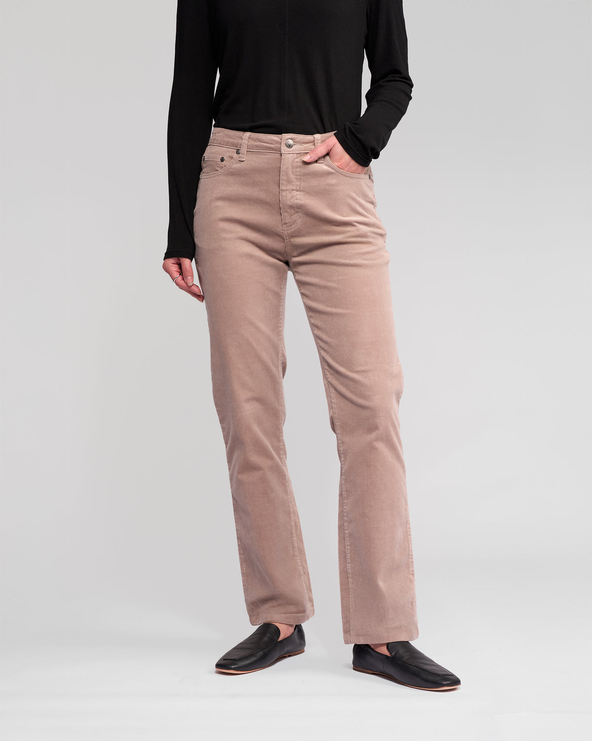 A person stands with one hand in their pocket against a plain light background, wearing the Vassalli Arch Cord Pant in Twig, a neutral beige straight-leg corduroy pant. Their head is not visible; they're also shown in a black long-sleeve top and black slip-on shoes.