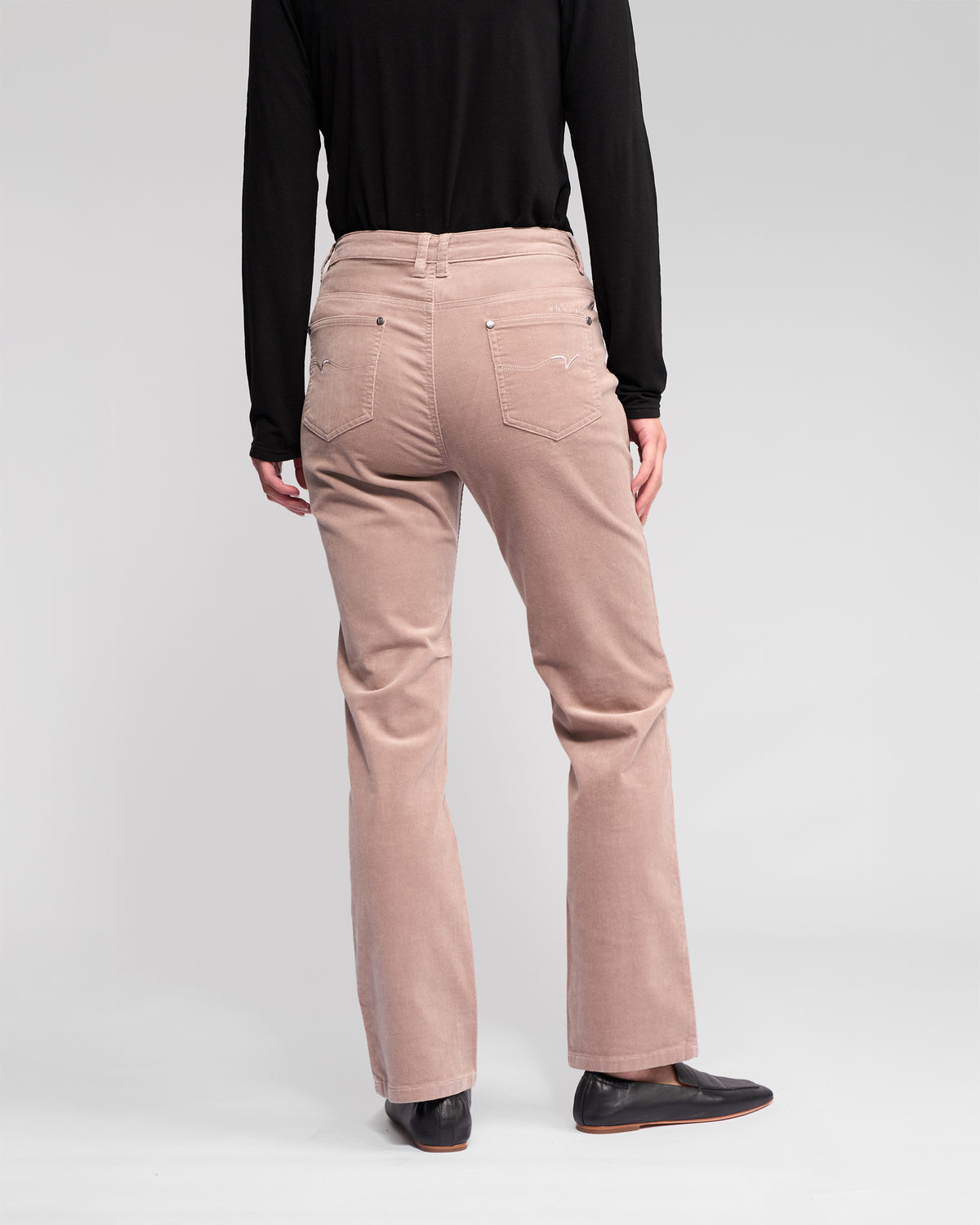 A person wearing a black long-sleeve top, Vassalli Arch Cord Pant in Twig (neutral beige), and black shoes stands facing away from the camera against a plain light background.
