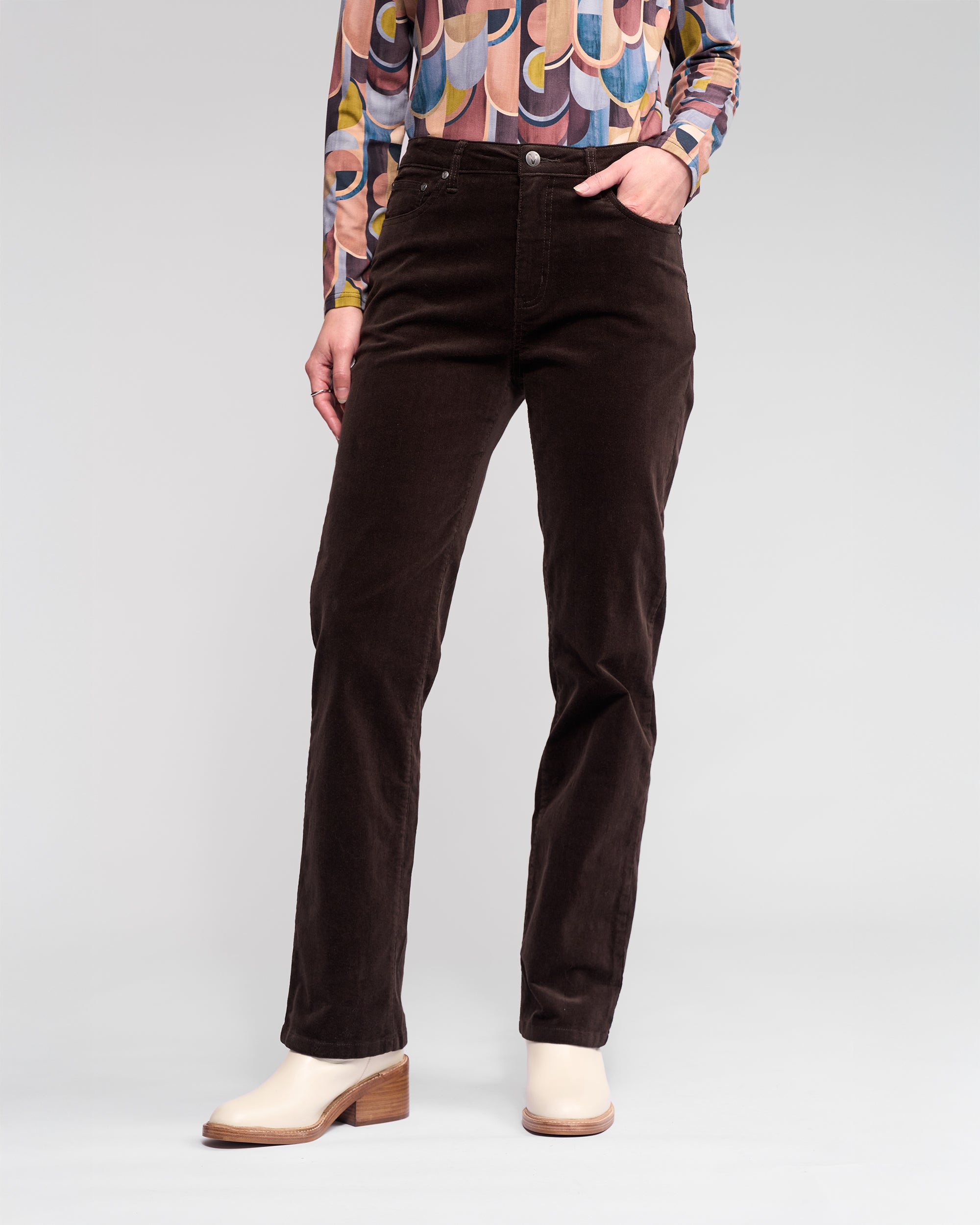A person models the Vassalli Arch Cord Pant in Walnut, paired with a colorful patterned long-sleeve shirt and light beige heeled shoes, standing against a plain background. Their face is not visible.