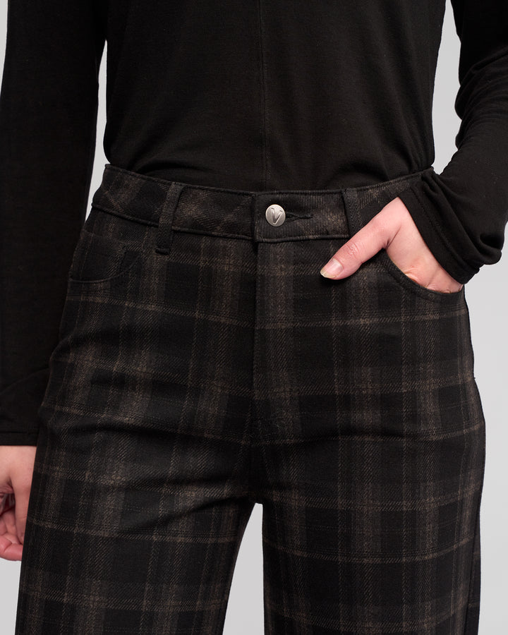 A person wears Vassalli's Empire Pant - Checkmate, with a black long-sleeve shirt, hand in pocket; only their torso and part of the legs are visible.