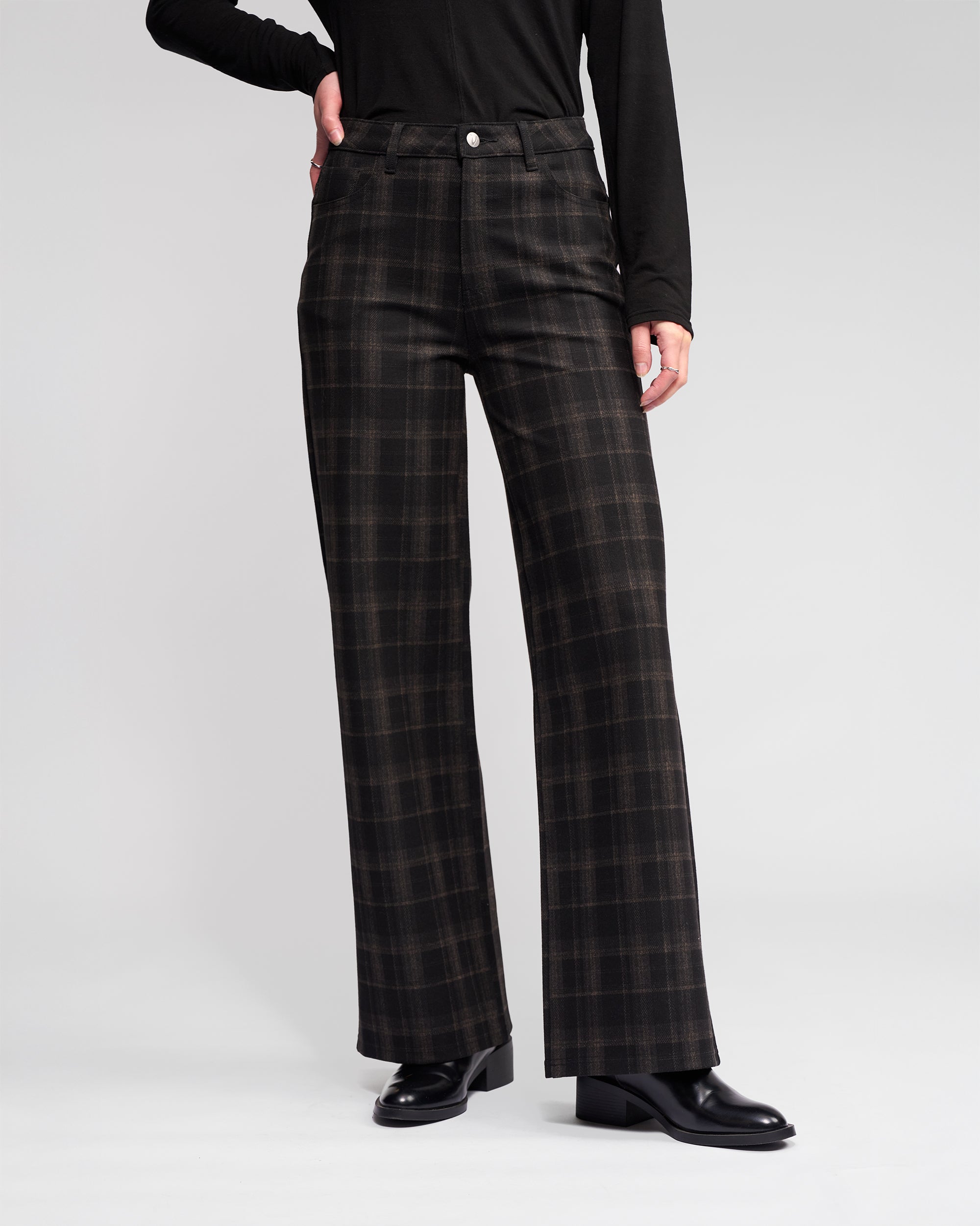 A person wears the Vassalli Empire Pant - Checkmate with a black long-sleeve top and black heeled shoes, standing against a light gray background—an effortlessly chic winter look.