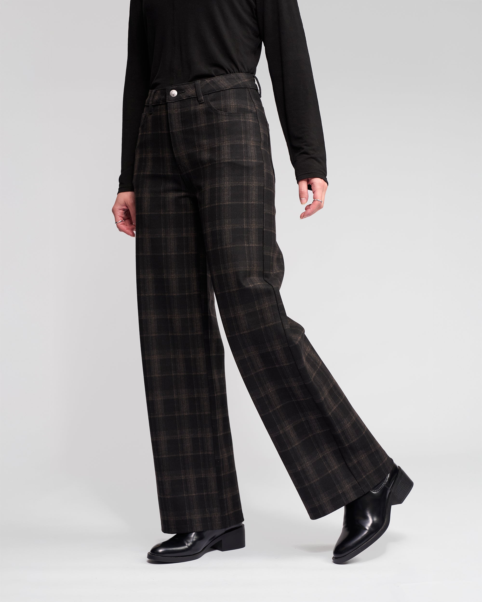 Model wears the Empire Pant - Checkmate by Vassalli: dark brown and black checkered wide-leg pants paired with a black long-sleeve top and black leather shoes, shown from the waist down against a plain light background—perfect winter style.