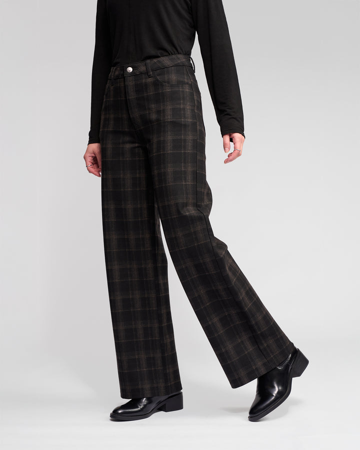 Model wears the Empire Pant - Checkmate by Vassalli: dark brown and black checkered wide-leg pants paired with a black long-sleeve top and black leather shoes, shown from the waist down against a plain light background—perfect winter style.