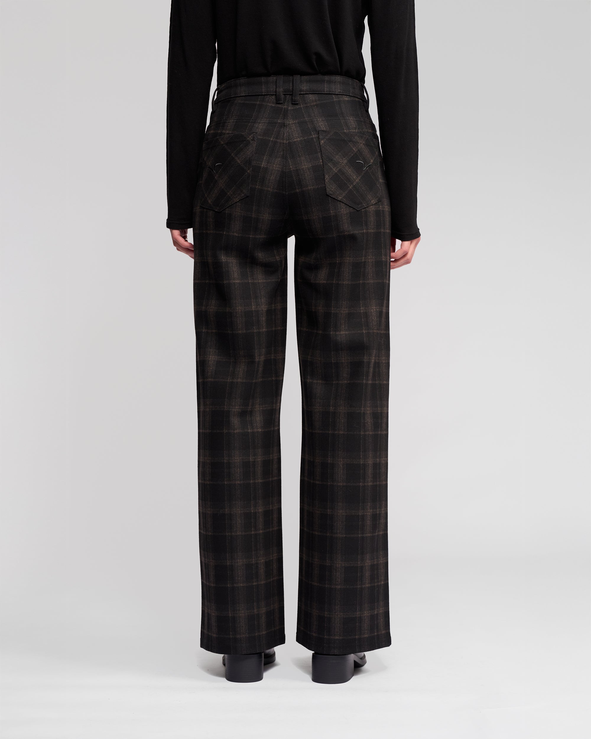 A person stands facing away, wearing Vassalli's Empire Pant - Checkmate (high-waisted, wide-leg checkered pants) with a black long-sleeve top and shoes—ideal winter wear against a light, plain background.