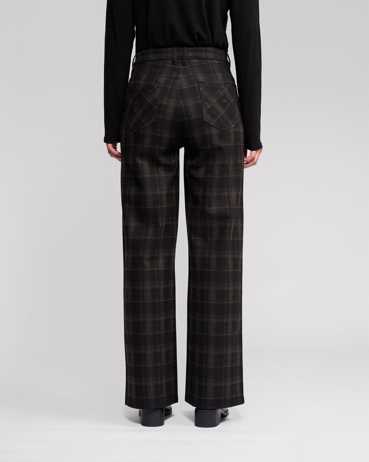 A person stands facing away, wearing Vassalli's Empire Pant - Checkmate (high-waisted, wide-leg checkered pants) with a black long-sleeve top and shoes—ideal winter wear against a light, plain background.