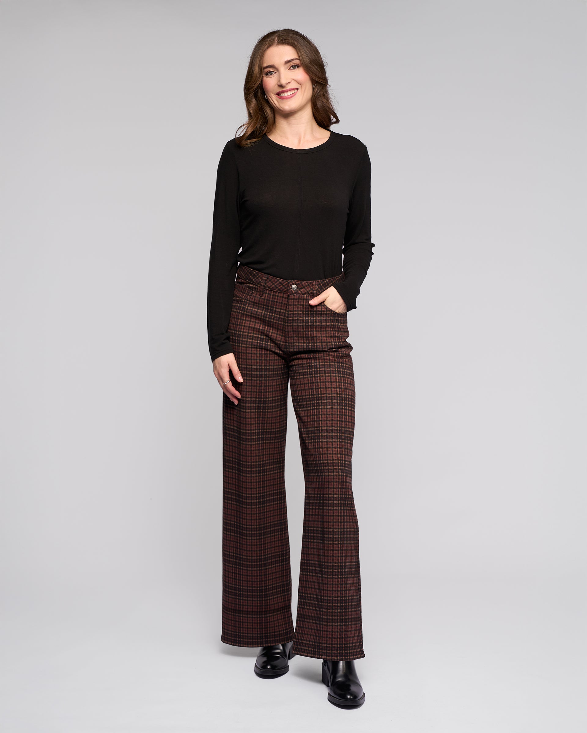 A woman stands against a plain background, smiling with one hand in her pocket. She wears a black long-sleeved top, Vassalli Empire Pant - Cherry—ideal for winter—and black shoes.