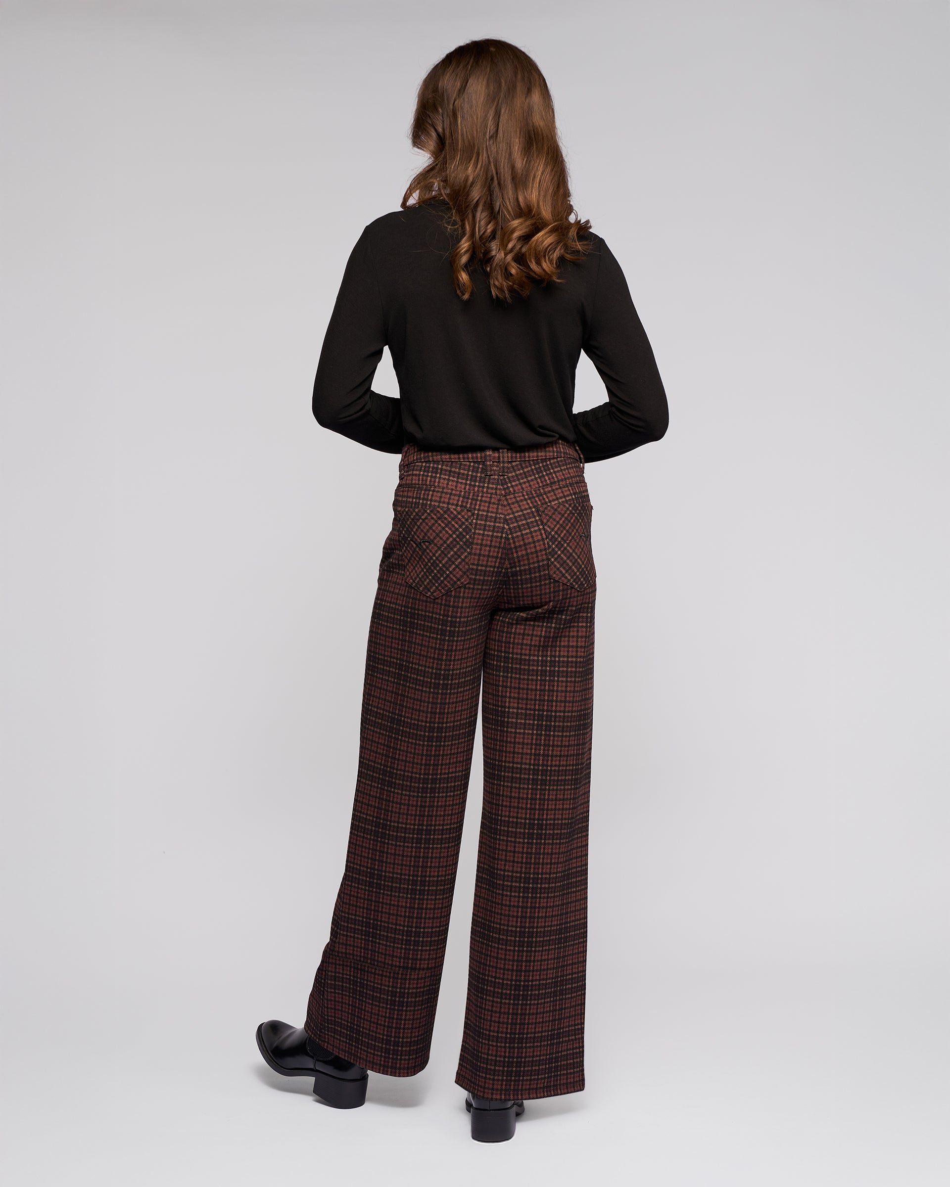 A person with wavy brown hair stands facing away, wearing a black long-sleeve top, Vassalli Empire Pant - Cherry in brown plaid, and black shoes, set against a plain light background.