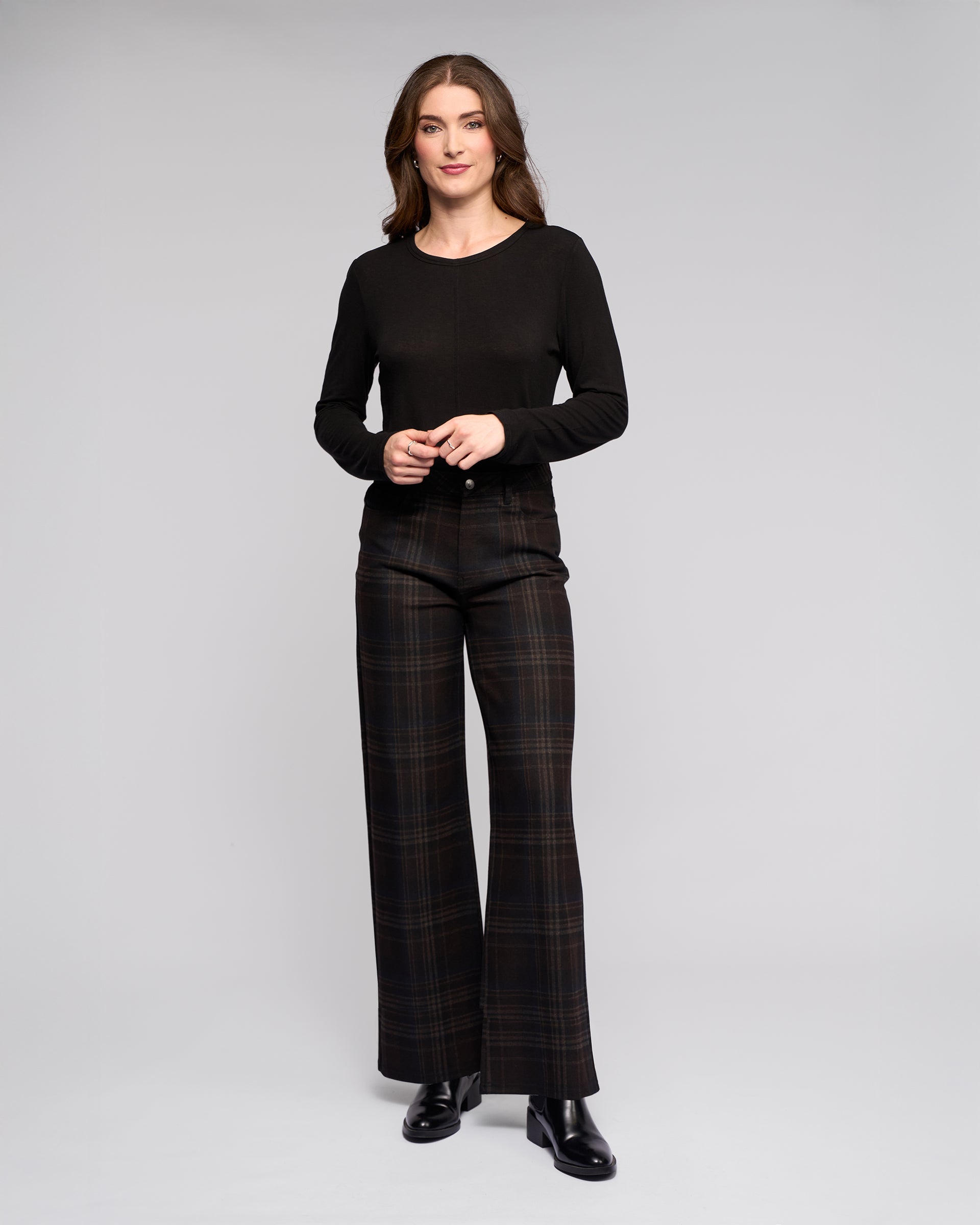 A woman smiles softly against a gray background, wearing a black long-sleeve top, Vassalli Empire Pant - Hugo high-waisted wide-leg pants, and black ankle boots for a chic winter look, with her hands lightly clasped.