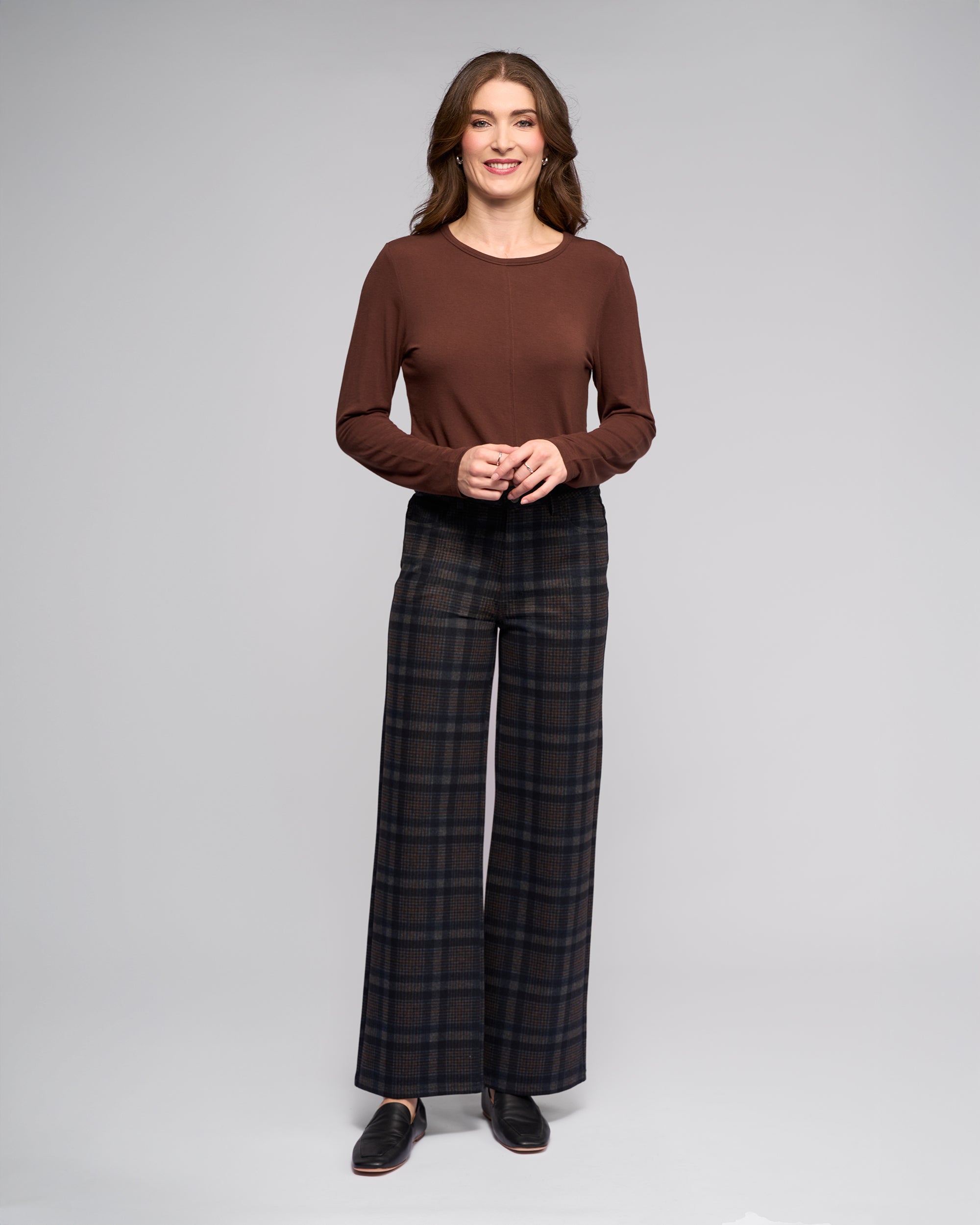 A woman with long wavy brown hair smiles against a plain background, wearing a Vassalli Empire Pant - Nomad in dark hues, a long-sleeve brown top, and black shoes. She gently clasps her hands in front of her.