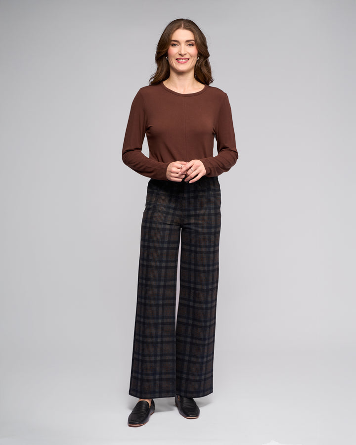 A woman with long wavy brown hair smiles against a plain background, wearing a Vassalli Empire Pant - Nomad in dark hues, a long-sleeve brown top, and black shoes. She gently clasps her hands in front of her.