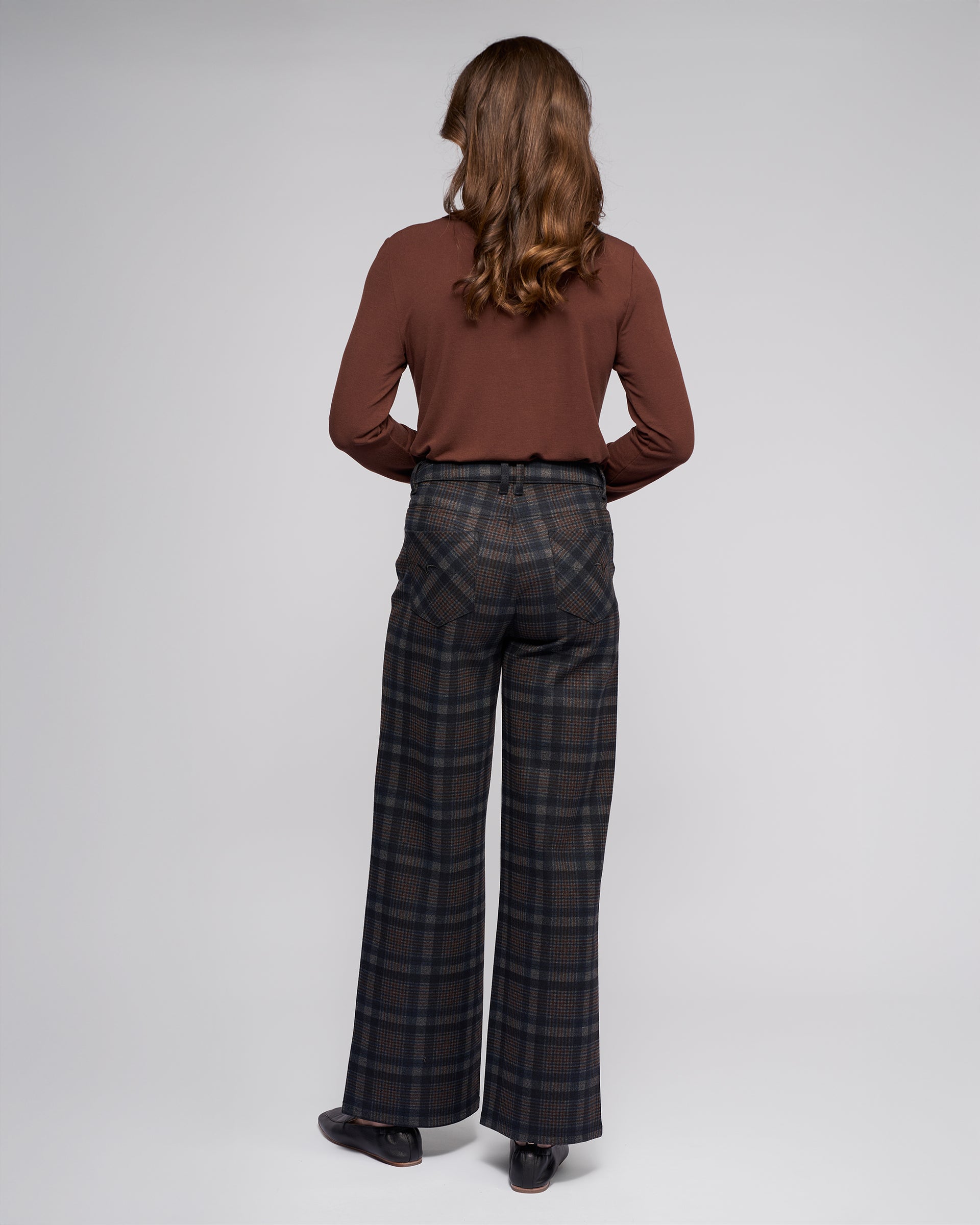 A person with long brown hair, facing away, wears a brown long-sleeve shirt and Vassalli’s Empire Pant - Nomad with dark shoes, standing against a plain light gray background.