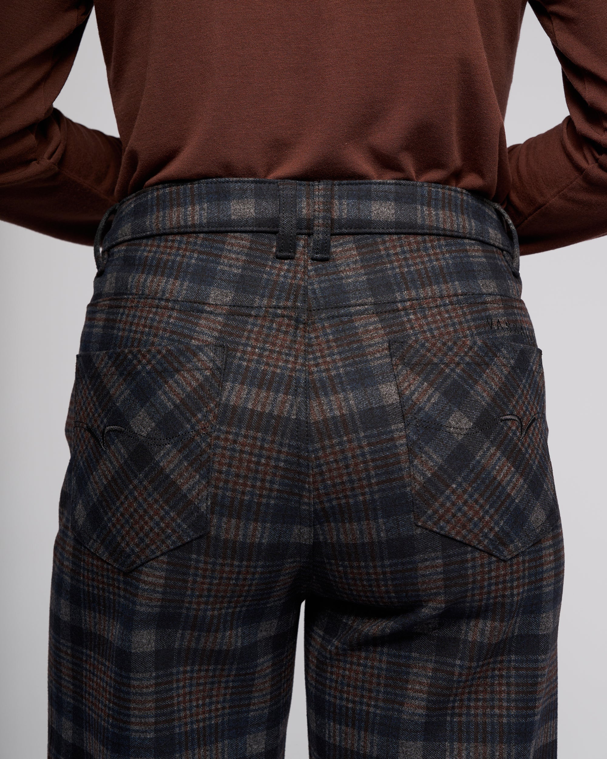 A close-up from behind shows a person wearing Vassalli’s Empire Pant - Nomad, high-waisted plaid pants in dark gray and brown, paired with a long-sleeve brown top. The photo highlights the pants’ pockets and belt loops—ideal for winter.