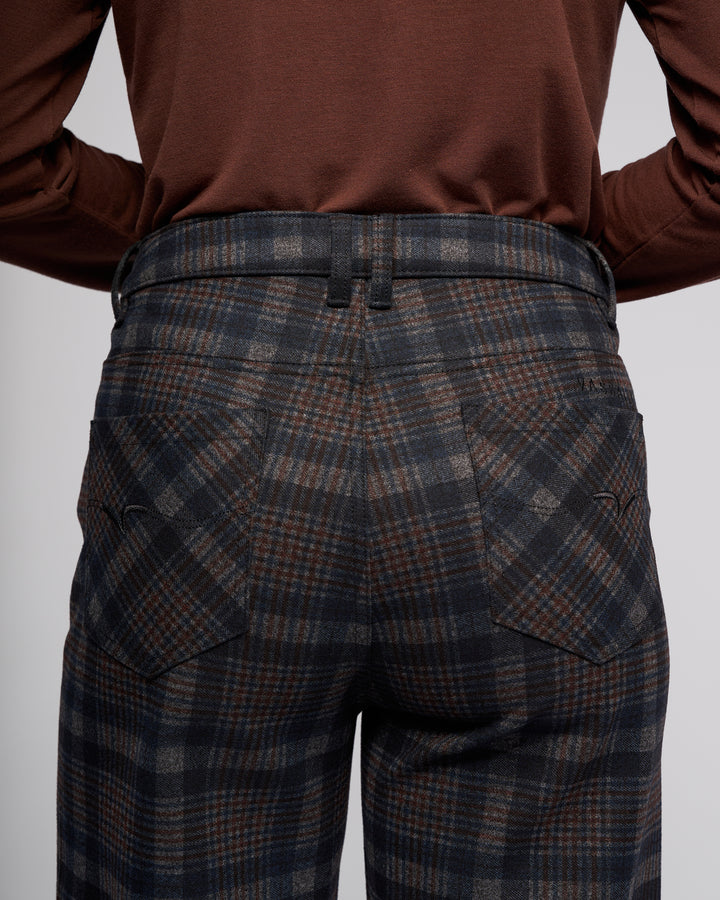 A close-up from behind shows a person wearing Vassalli’s Empire Pant - Nomad, high-waisted plaid pants in dark gray and brown, paired with a long-sleeve brown top. The photo highlights the pants’ pockets and belt loops—ideal for winter.
